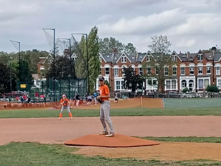 A youth pitcher in an orange Fall Ball shirt on the mound at Finsbury Park, with a fielder behind him and the Endymion Road houses beyond